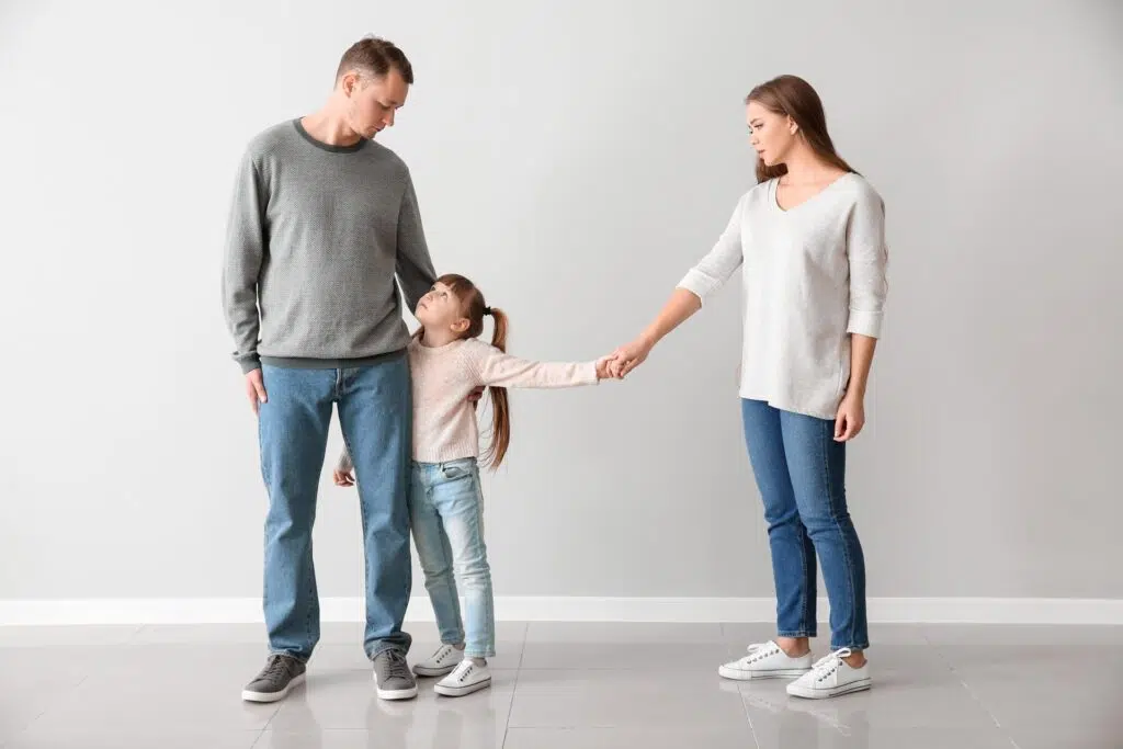 A father and mother stand next to each other with their daughter breaking free of the mom's hand and wrapping her hand around her father.