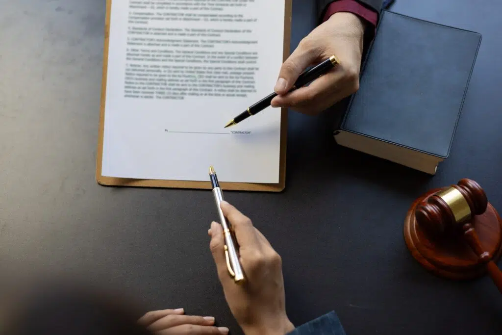 Two hands holding pens pointing at a contract on a desk next to a gavel.