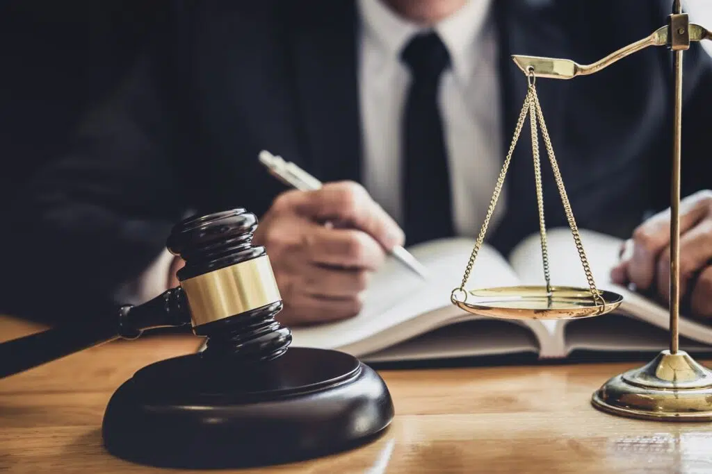 A man in a suite writing in a notebook with a pen in front of a gavel and scales of justice.
