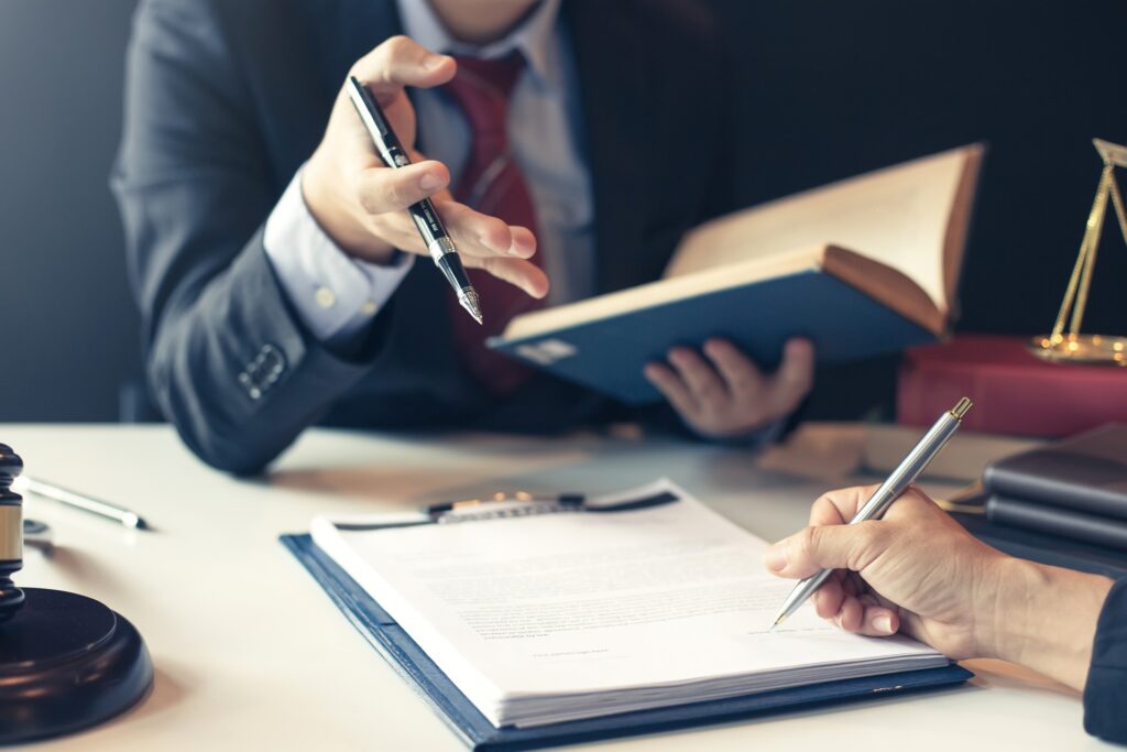 A man sits at a desk holding a pen and book while talking to a woman writing on a sheet of paper on a clipboard.