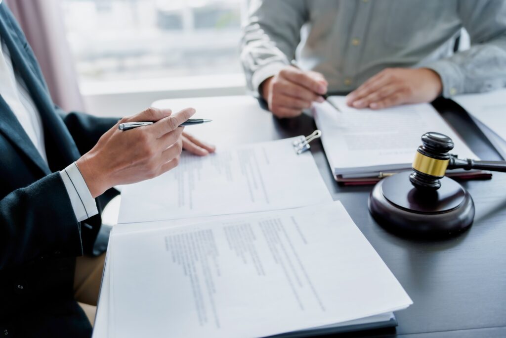 A man sits with an attorney at a desk signing papers next to a gavel resting on its stand.