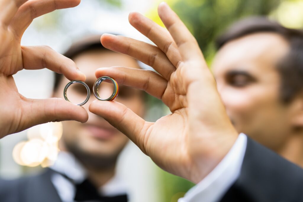Two men in suits hold their wedding rings up next to each other.