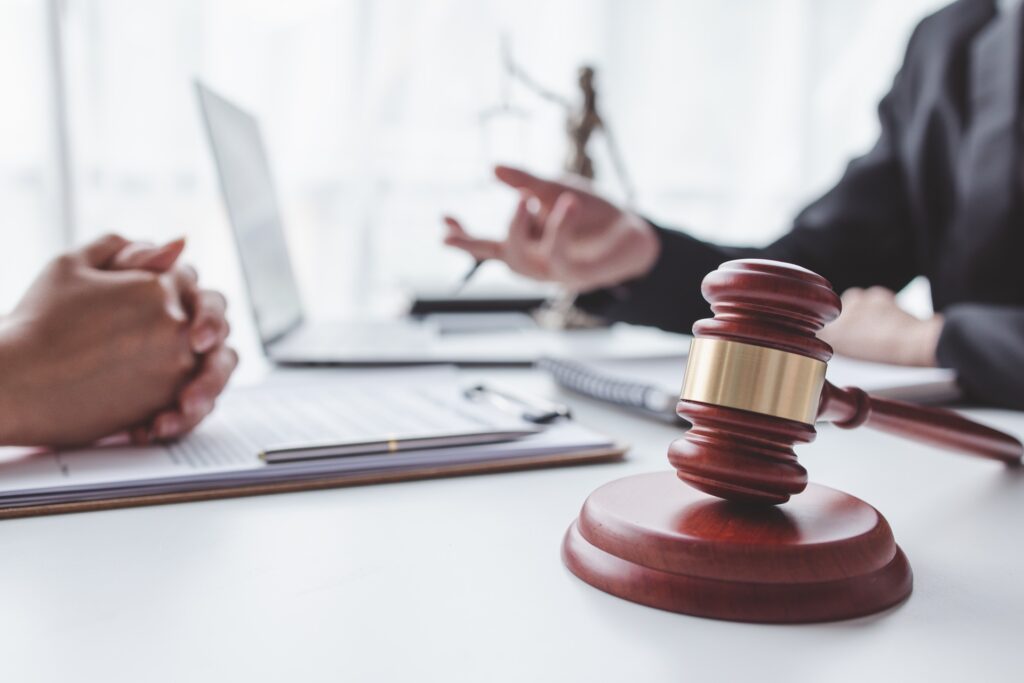 An attorney sits across a desk from a client talking in front of a laptop and a gavel resting on its stand.