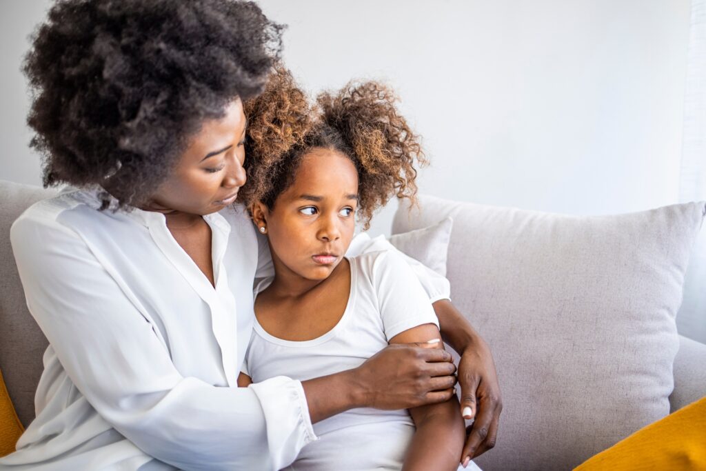 A woman sitting on a couch with her arms clasped around her daughter who looks upset.
