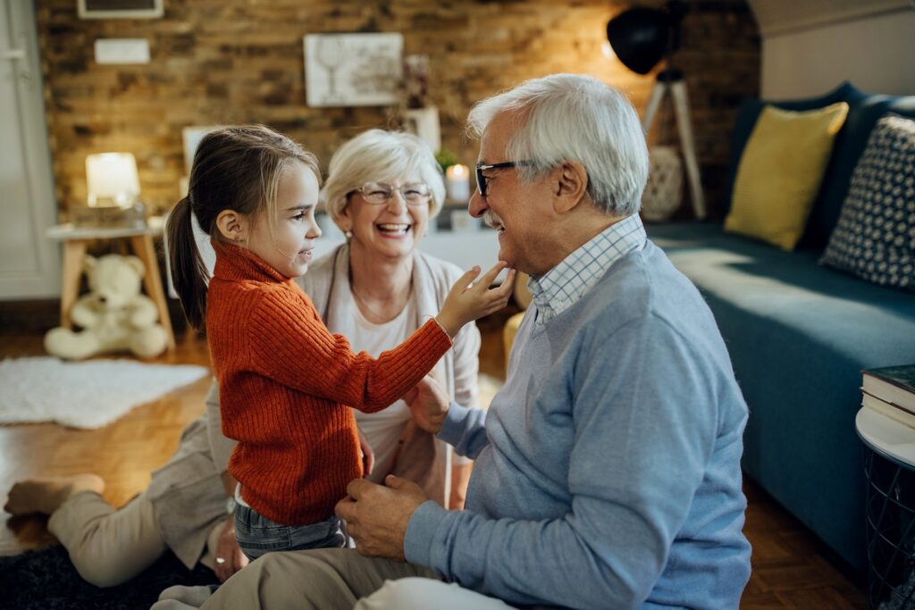 A grandmother and grandfather sitting on the ground with their granddaughter in a living room smiling.