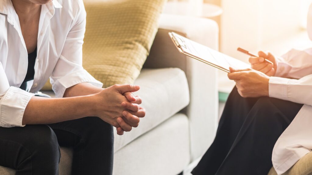 Two adult females sit across from each other on a couch and chair talking while one holds a clipboard.