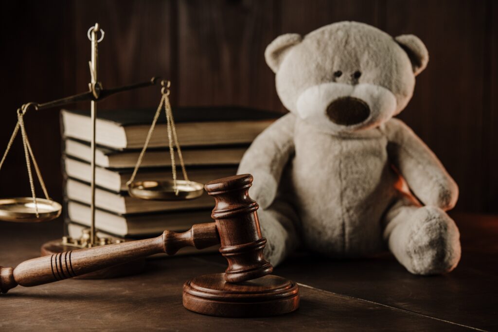 A teddy bear sits on a desk in front of a stack of law books, scales of justice, and a gavel resting on its stand.