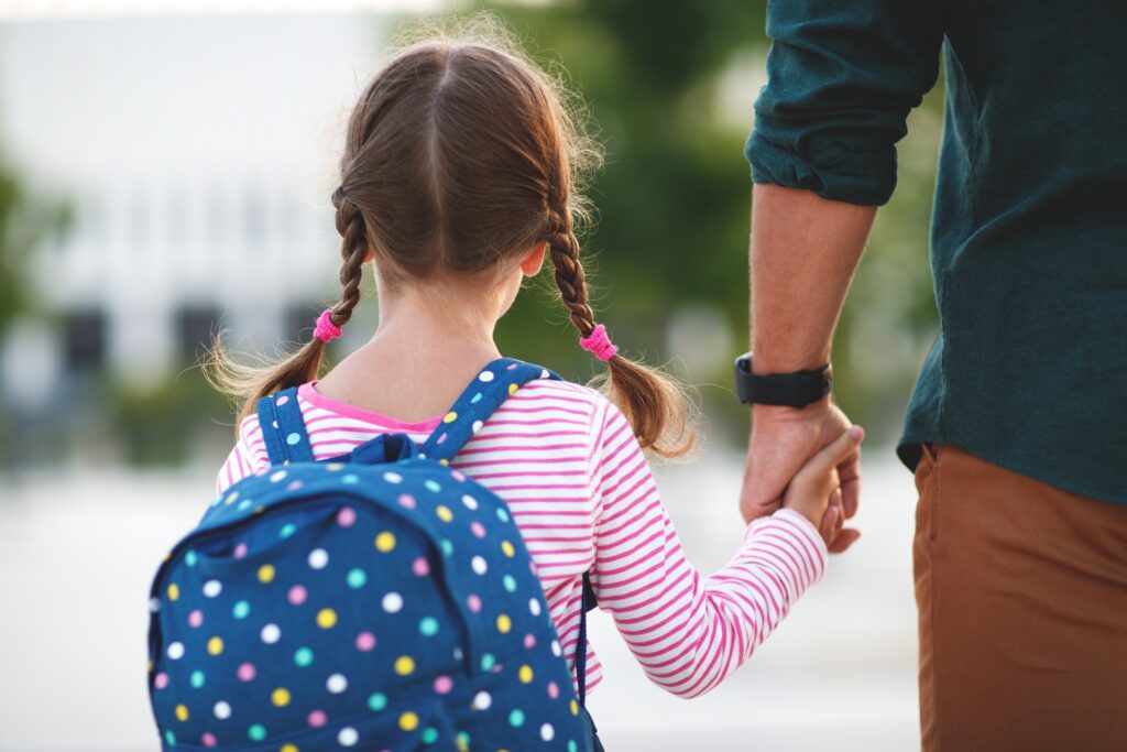 A female child wearing a blue backpack holding her fathers hand walking on a sidewalk.