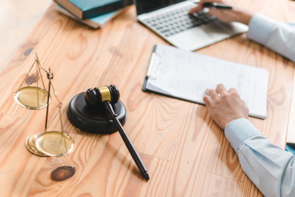 A lawyer sits at a desk with his hand resting on a clipboard and a silver laptop next to a gavel resting on its stand and scales of justice.