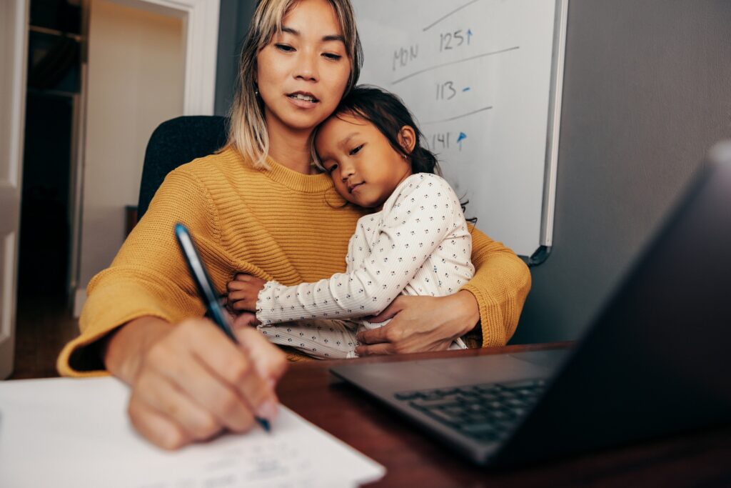 A woman holds her daughter in her lap while writing on a paper next to her laptop.