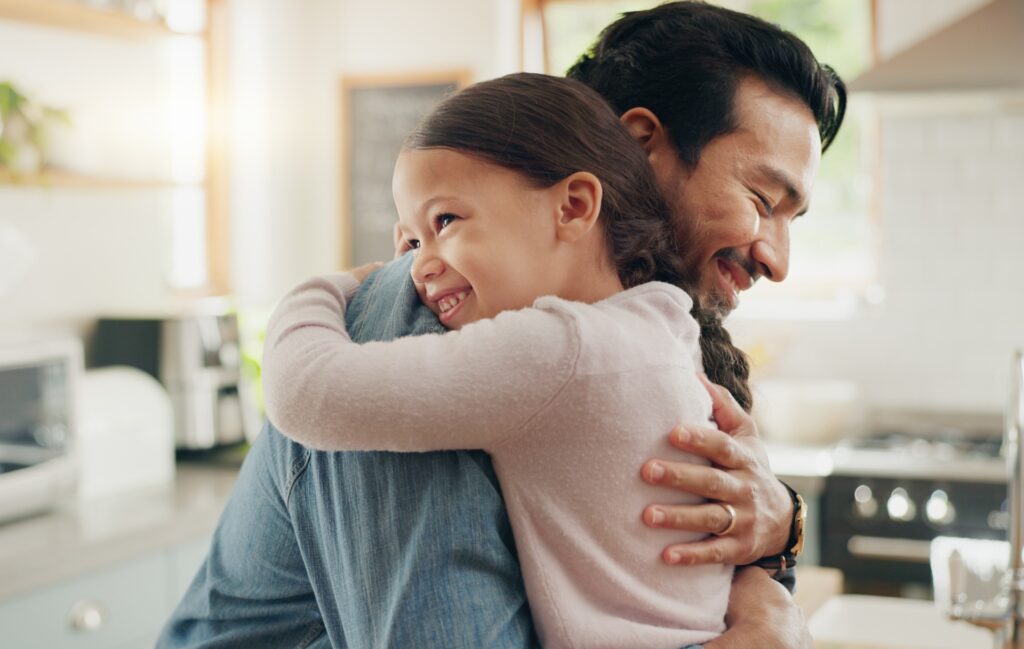 A man hugs his daughter while smiling inside a house.