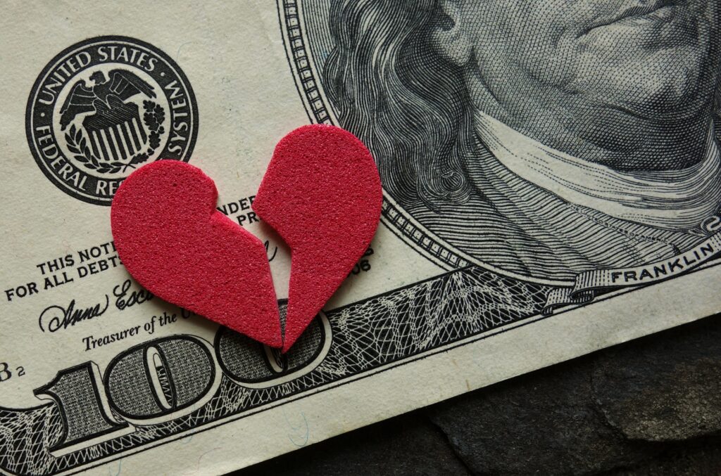 A broken felt heart sits on a hundred dollar bill.