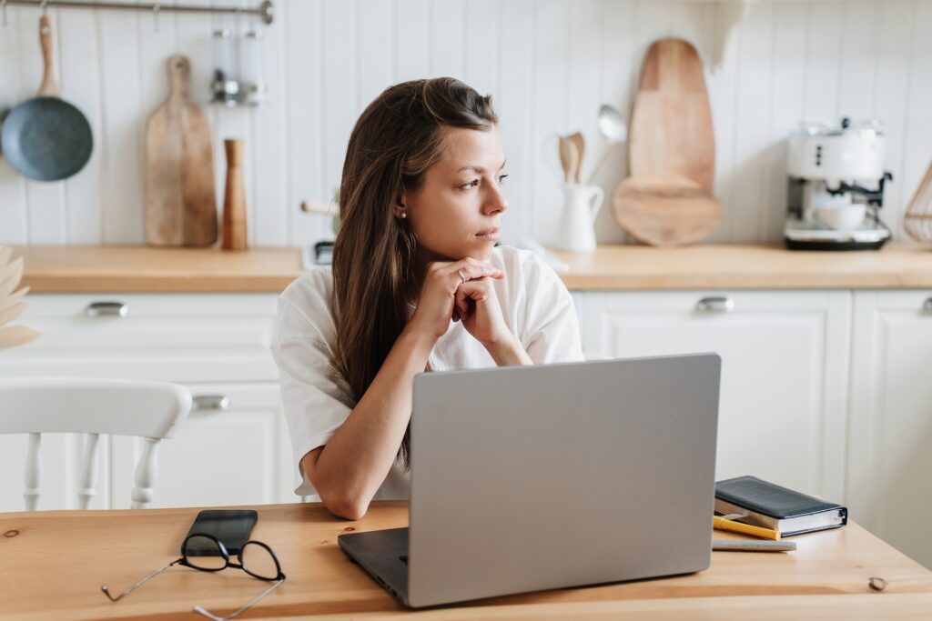 A woman sitting at a table in front of a silver laptop sits with her hands clasped below her chin looking to the side deep in thought.