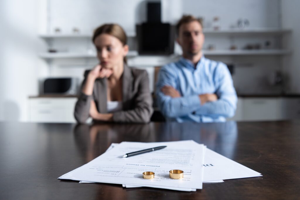 A couple sits next to each other at a table in front of divorce papers with wedding rings and a pen resting on them.