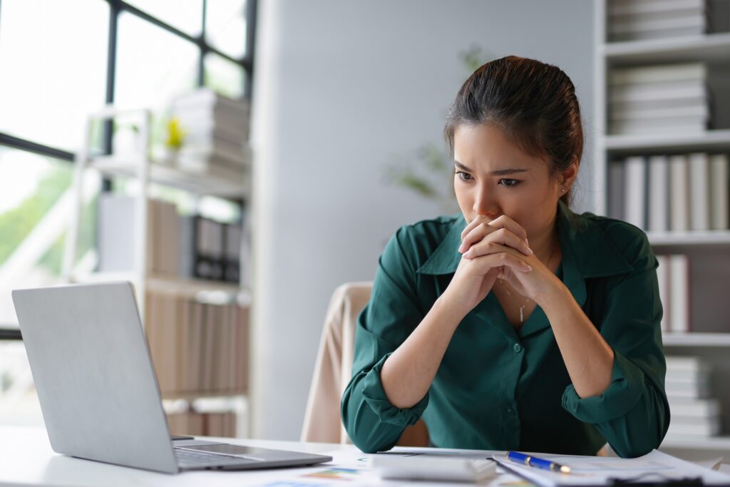 A woman sitting at a desk behind a silver laptop with her hands clasped in front of her mouth looking concerned.