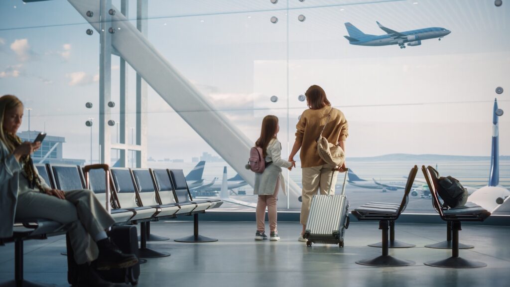 A woman holds hands with her daughter looking out the window of an airport terminal.