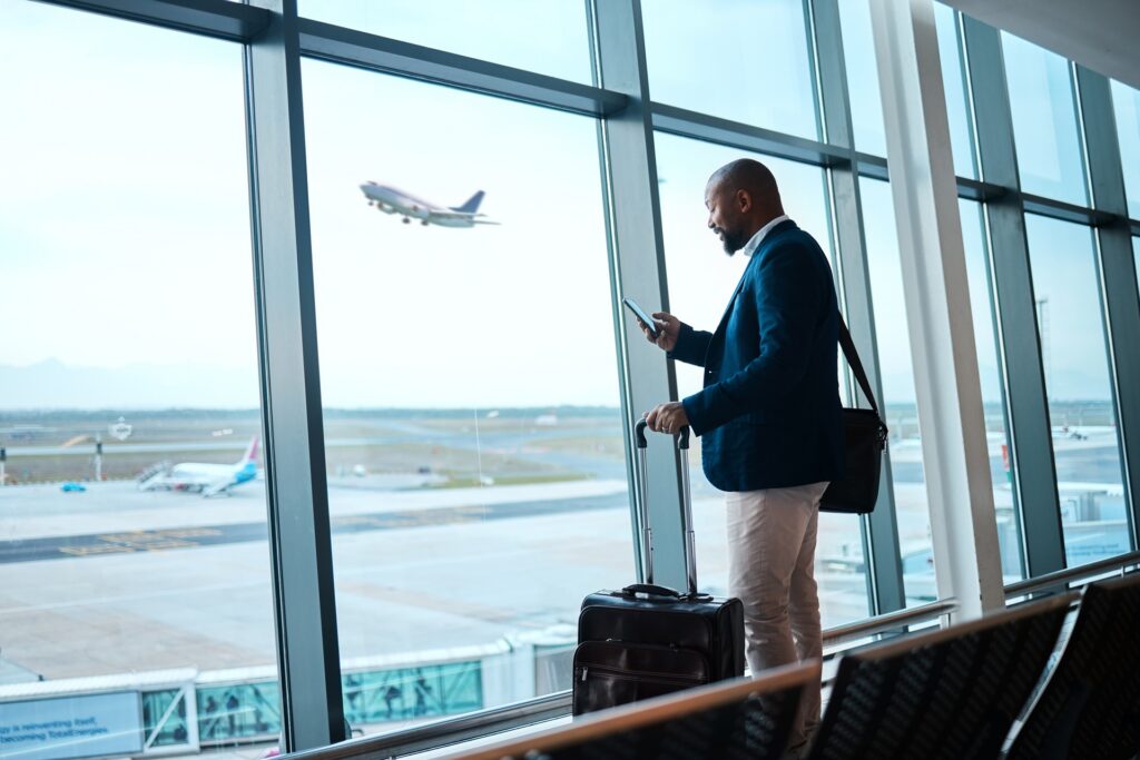 A person looks at their cell phone with luggage near them standing near a window in an airport with a plane taking off in the distance.
