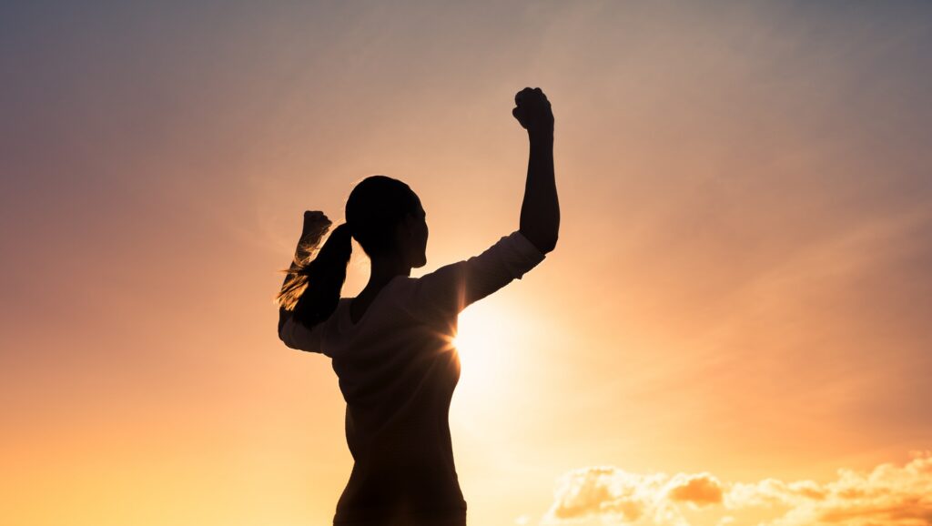 The silhouette of a person holding their arms in their air against a sunny backdrop.