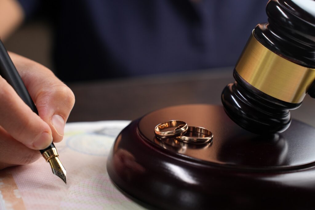 A person writing with a pen on a sheet of paper next to two wedding rings sitting next to a gavel on its stand.