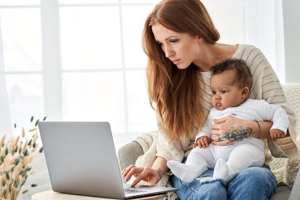 A woman holds her child while looking at a laptop with a serious expression.