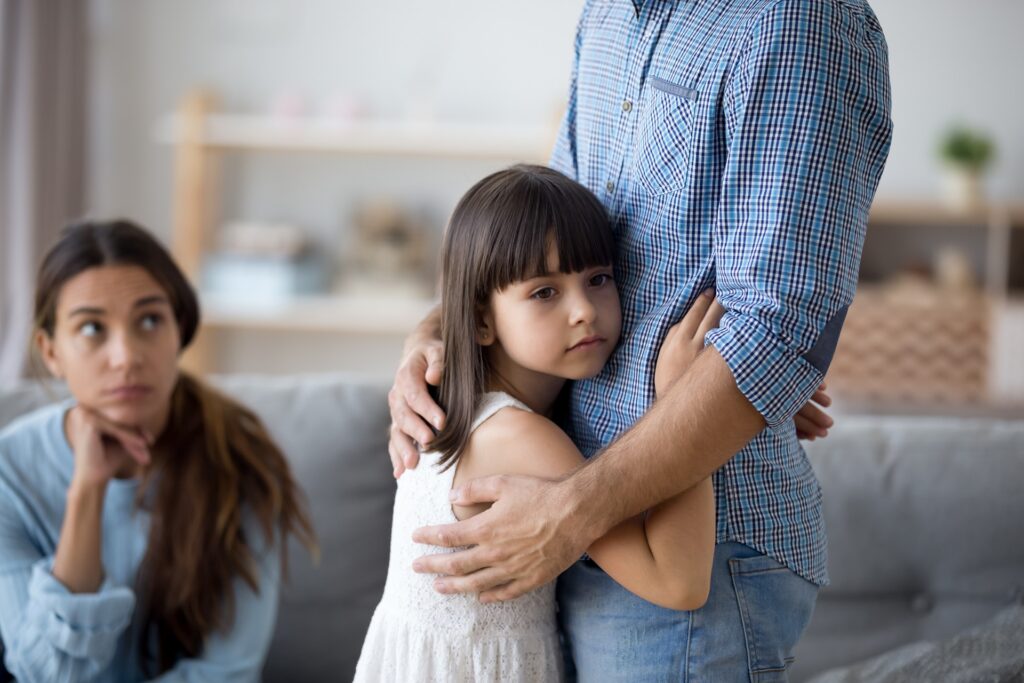 A young girl hugs her father as her mother sits on a couch in the background.