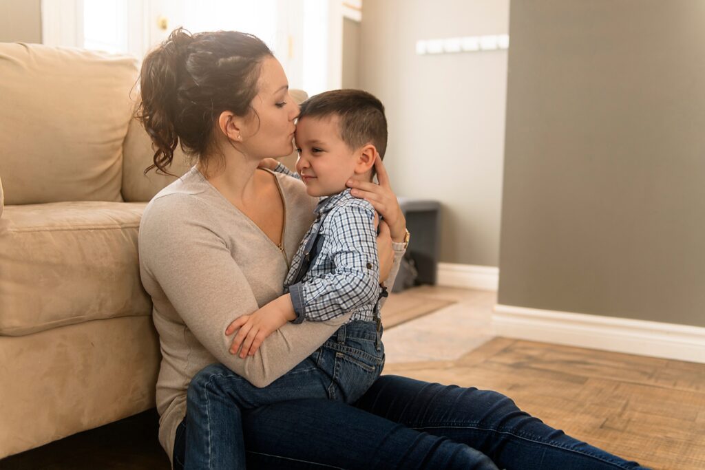 A woman kisses her sin on the forehead while sitting down in front of a couch on the ground.