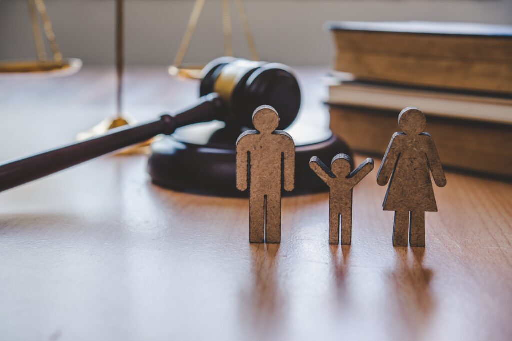 A wooden cutout of a family sitting on a wooden desk in front of books, scales of justice, and a gavel resting on its stand.