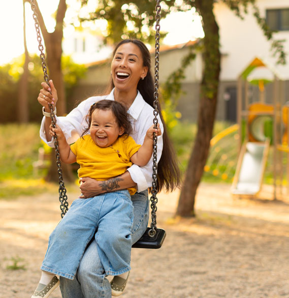 Mother and young child smiling together on a playground swing | Hemmat Law Group