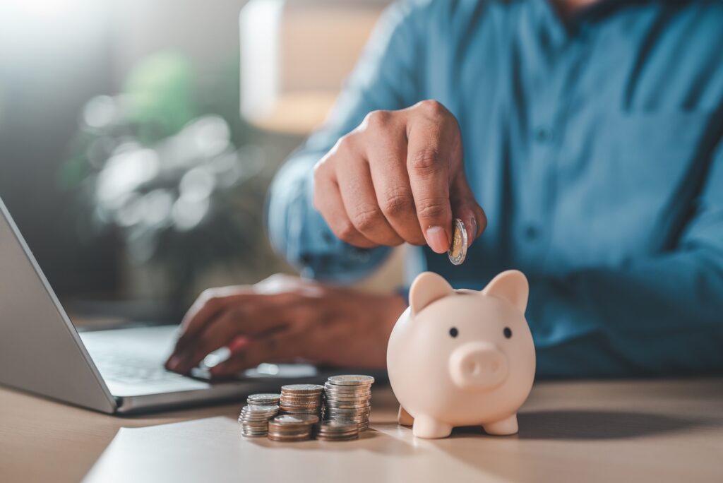 Person adding coins to piggy bank beside laptop & savings | Hemmat Law Group